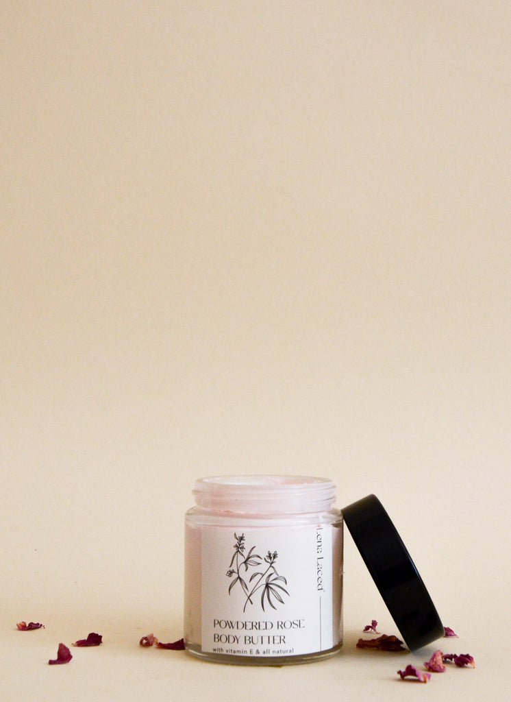 A 4 oz jar of Lena Laced, "Powdered Rose Body butter." The product is a light pastel pink and the jar is surrounded by rose petals.  